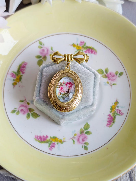 Gold locket brooch with vintage pink rose on blue floral design displayed on a decorative plate