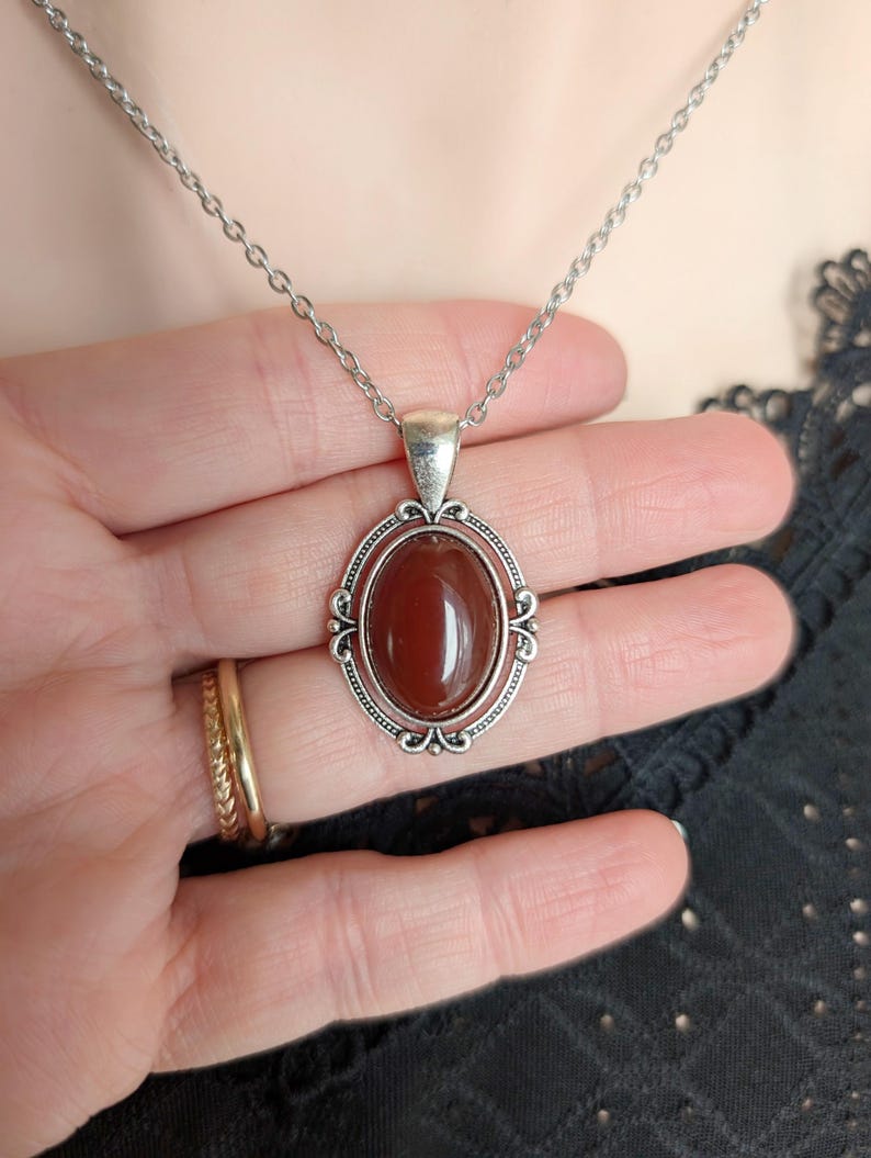 Hand holding a Blood Red Agate Crystal Necklace - Silver Flourish Pendant Setting against a neutral background.