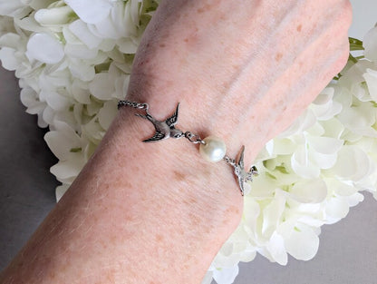 Hand wearing a Silver Swallow Bracelet with Glass Pearl and Toggle Clasp against a white floral background