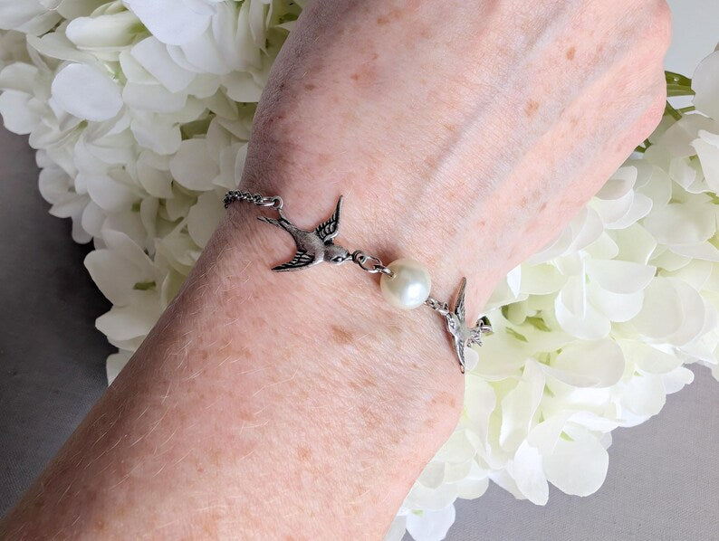 Hand wearing a Silver Swallow Bracelet with Glass Pearl and Toggle Clasp against a white floral background