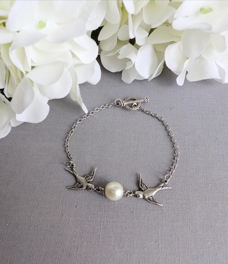 Silver Swallow Bracelet: Glass Pearl Bird Jewelry with Toggle Clasp on a gray surface with white flowers in the background