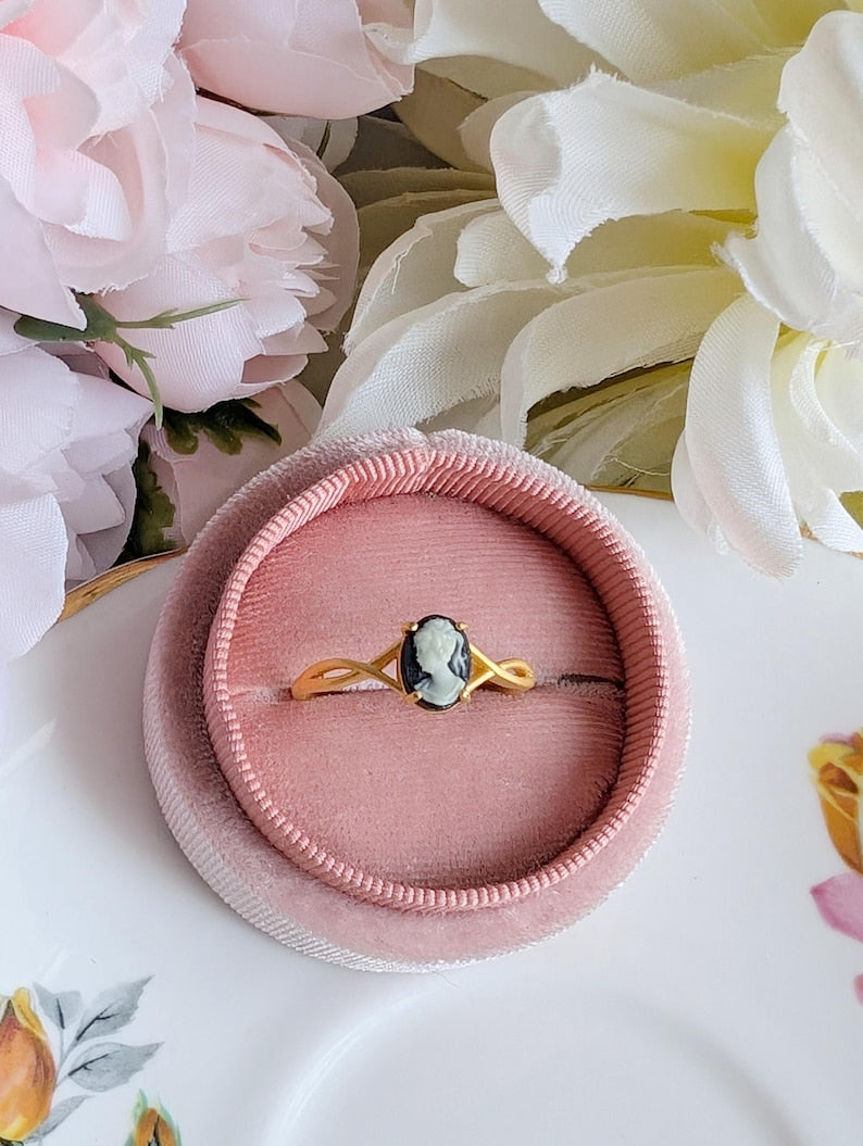 Matte gold cameo ring with single loop shoulder detail and black cameo in a pink velvet box surrounded by flowers
