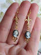 Gold fleur-de-lis earrings with cameo stones held between fingers against a light background