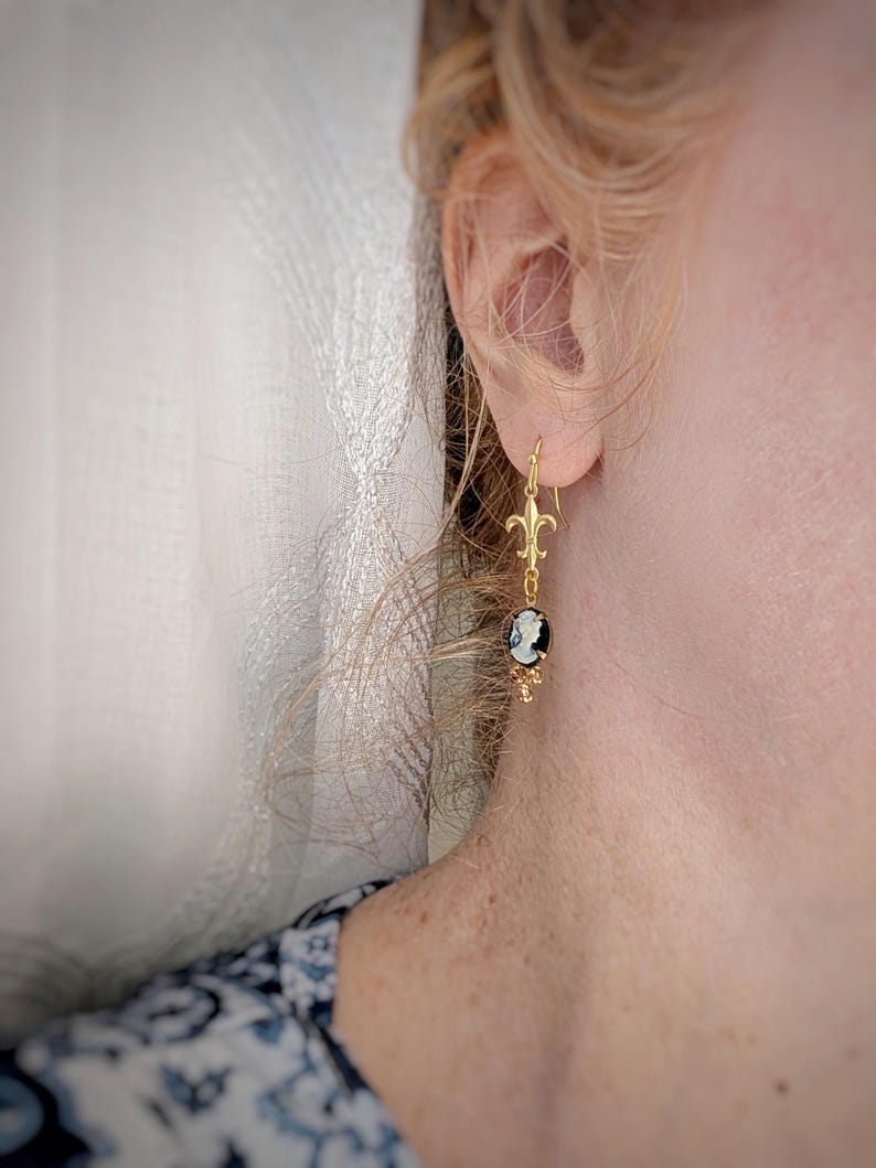 Close-up of an ivory on black cameo earring with gold Fleur de Lis connector and cognac Austrian crystals worn by a person.