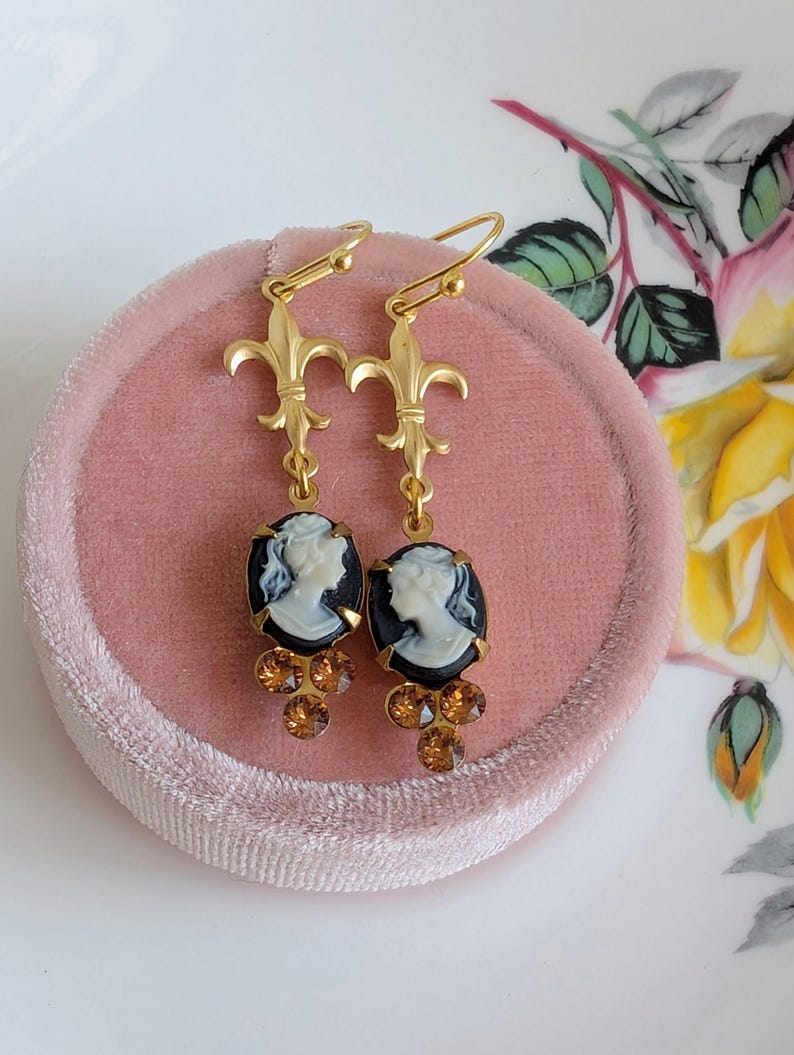 Handmade ivory on black cameo earrings with gold Fleur de Lis connectors and cognac Austrian crystals on a pink jewelry box with floral background.