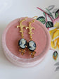 Handmade ivory on black cameo earrings with gold Fleur de Lis connectors and cognac Austrian crystals on a pink jewelry box with floral background.