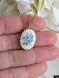 Blue Rose Vintage Porcelain Necklace - 1980s Limoges Style - Brass Setting held between fingers against a floral background