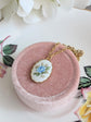 Blue Rose Vintage Porcelain Necklace - 1980s Limoges Style on a pink velvet cushion, floral background