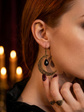 A close-up lifestyle shot of a woman with red hair wearing an ornate bronze crescent moon dangle earring with a black teardrop pendant, posed in front of warm, glowing candles.